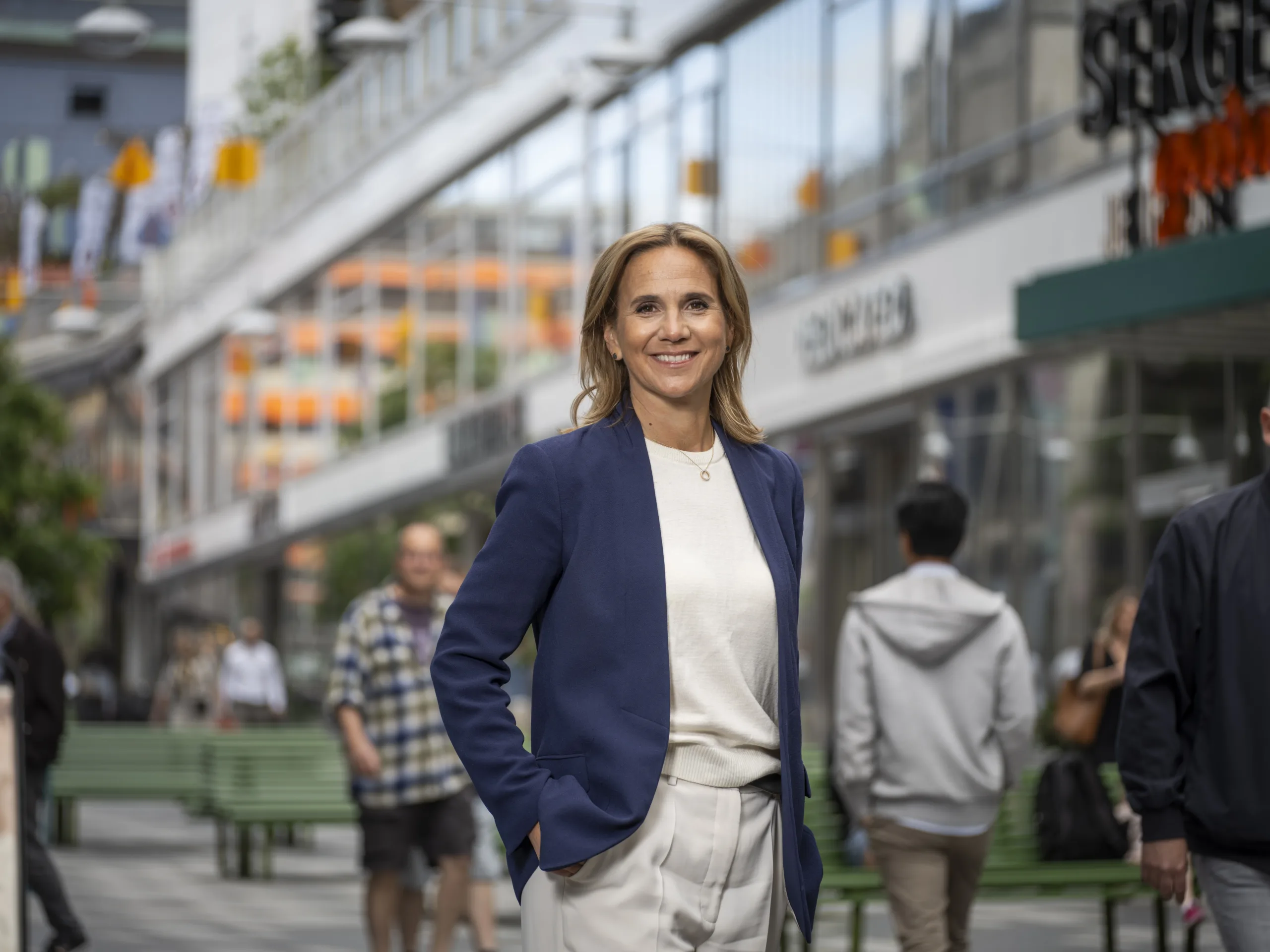 En kvinna i blå kavaj och vit blus står leende på en stadsgata med människor och butiker i bakgrunden.