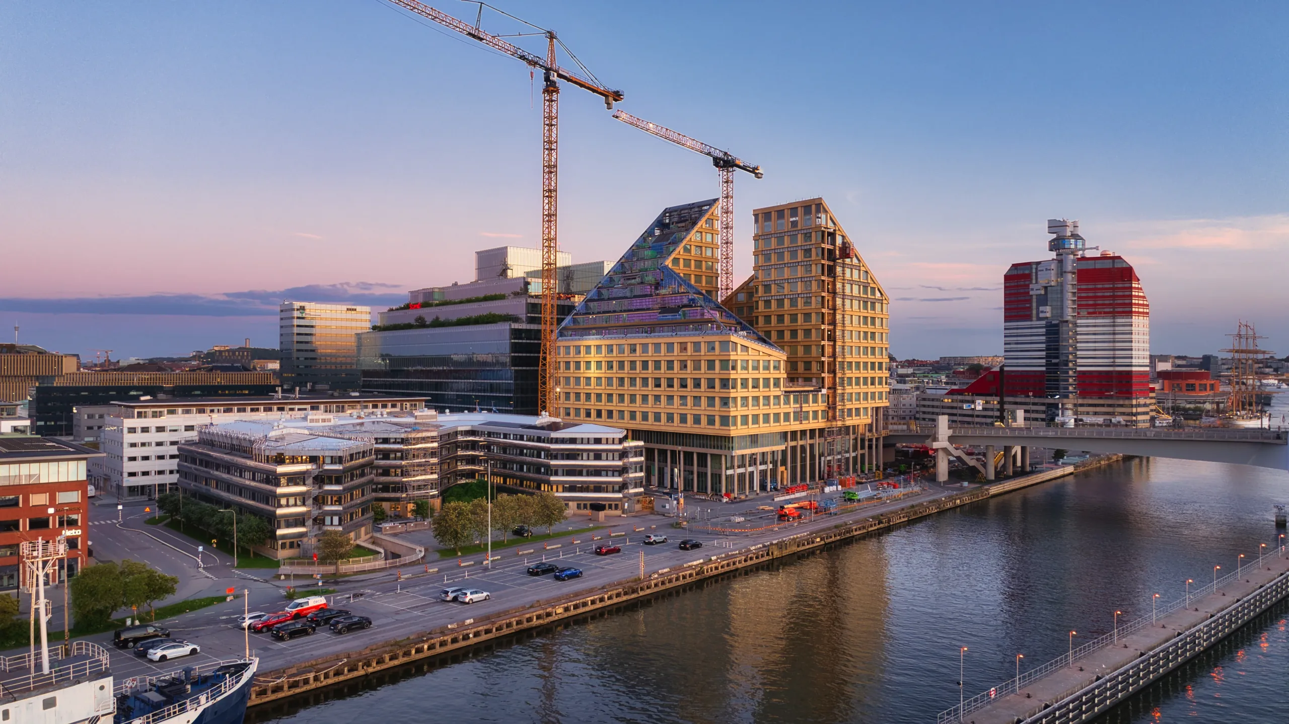 En byggarbetsplats vid vattnet med moderna byggnader, lyftkranar och en väg med bilar.