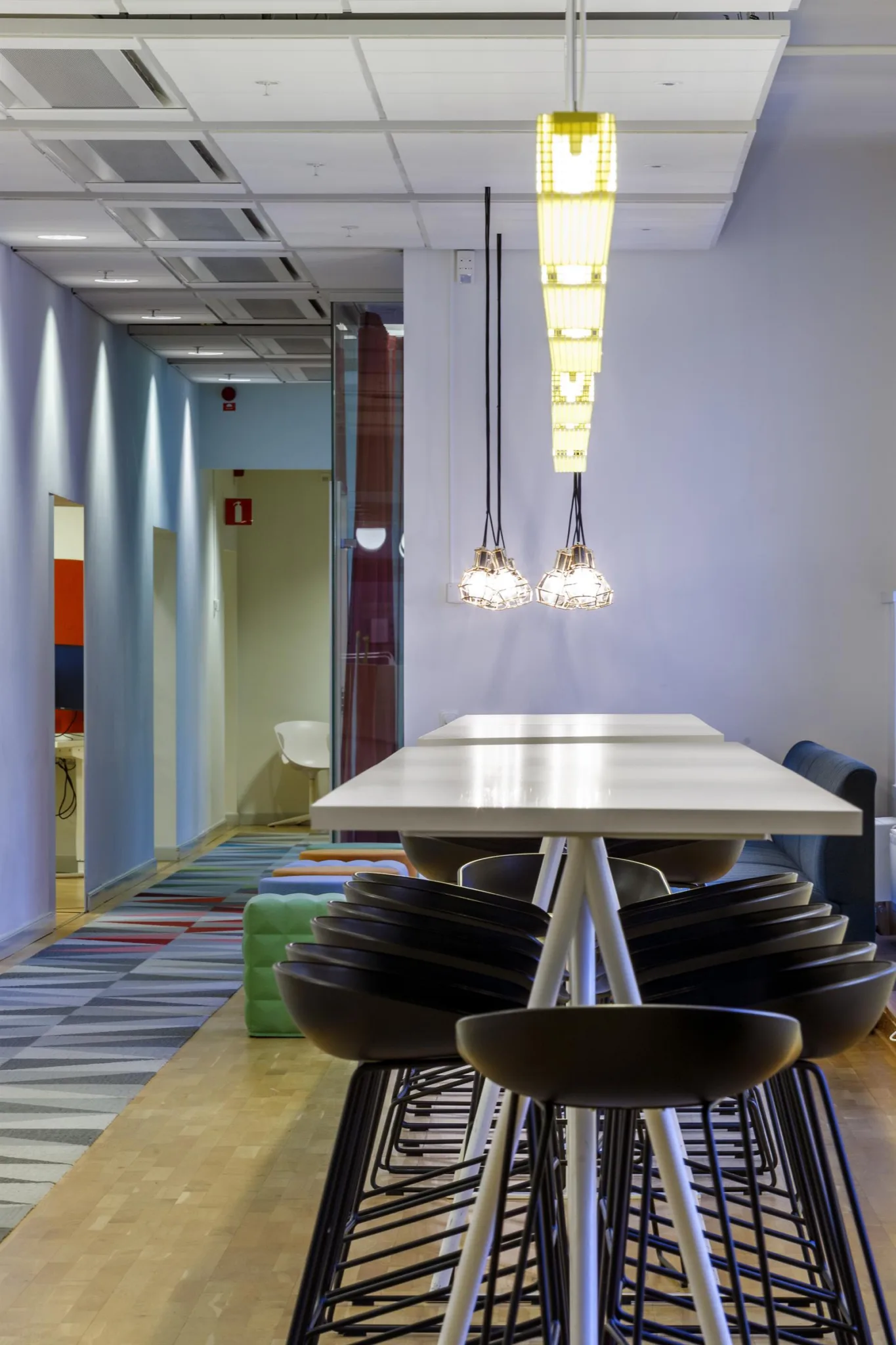 5. En lounge med lång bord, svarta stolar, taklampor och en färgglad matta som leder till en korridor.