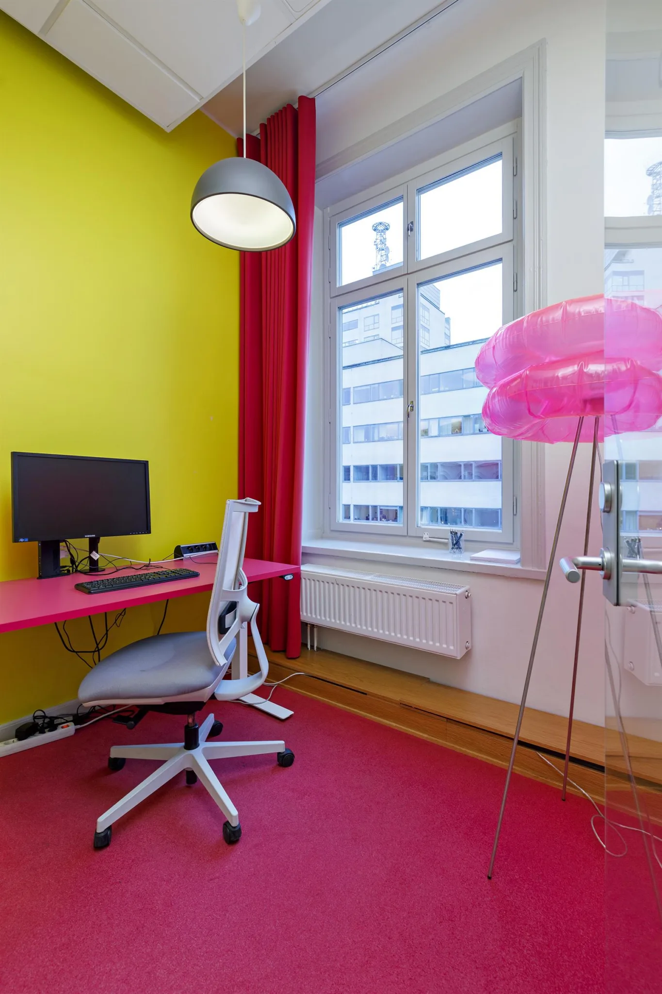 Ett färgglatt kontorsrum med gul vägg, rosa skrivbord, vit stol och en rosa lampa.