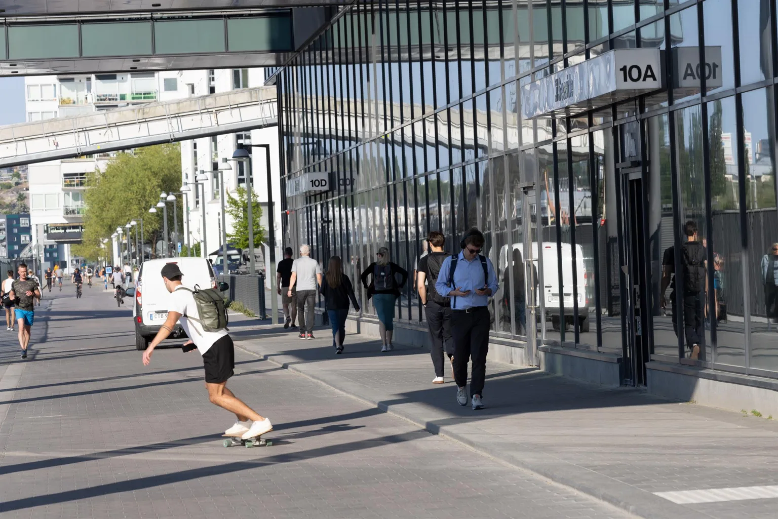 En trottoar utanför en glasfasad byggnad där människor går och en person åker skateboard.