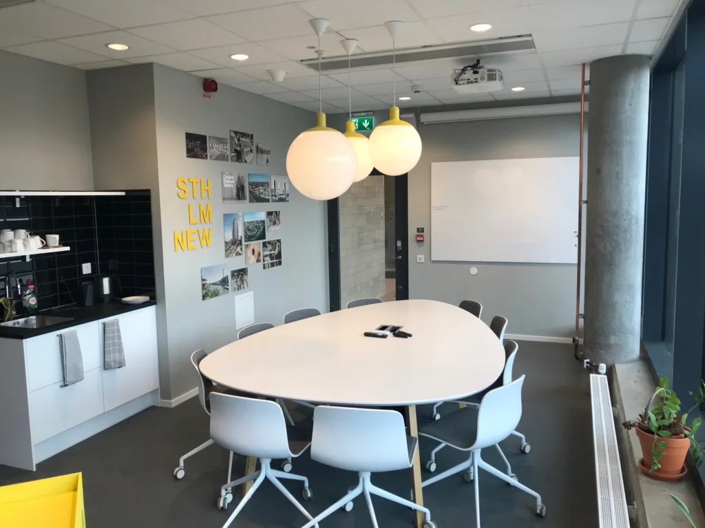 5. Ett mötesrum med ett ovalt bord, vita stolar, en whiteboard och dekorativa lampor.