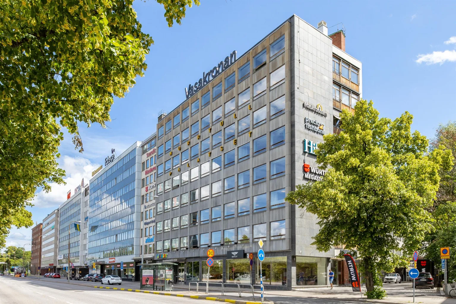 En modern byggnad med glasfasad och flera våningar, omgiven av träd och en gata.