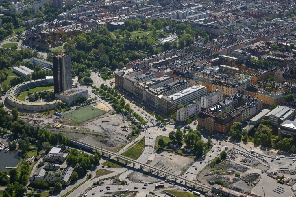 Flygfoto över en stadsdel med byggnader, vägar och grönområde i bakgrunden.