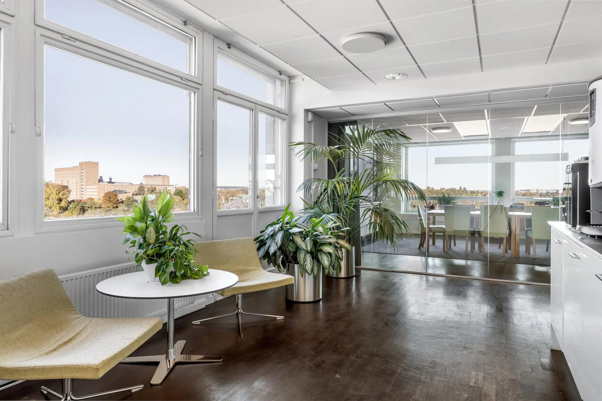 Ett modernt väntrum med stora fönster, växter, sittplatser och en öppen planlösning mot en kaffestation.
