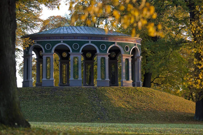 4. En rund paviljong med pelare står på en gräskulle omgiven av höstfärgade träd.