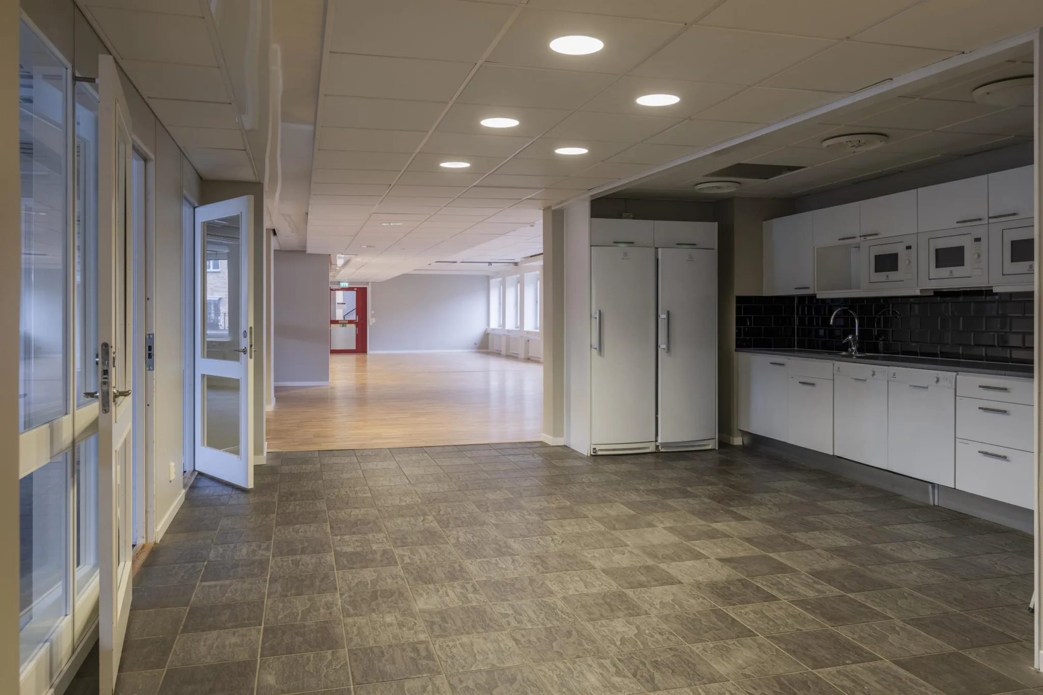 Interiör av en byggnad med klinkergolv, köksutrustning till höger och en öppen korridor som leder till ett ljust utrymme.