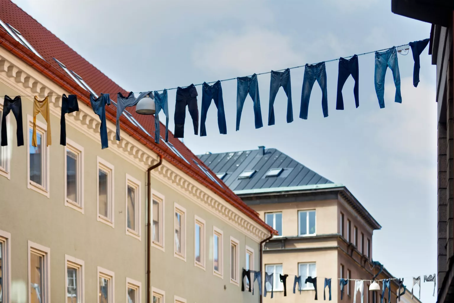 Jeans hänger på tvättlinor mellan byggnader med ljusa fasader under en blå himmel.
