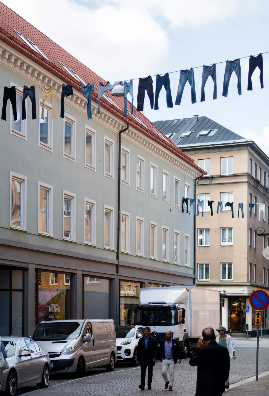 Byggnader med ljusgrön fasad, där jeans hänger på tvättlinor ovanför gatan med skyltfönster och parkerade fordon.