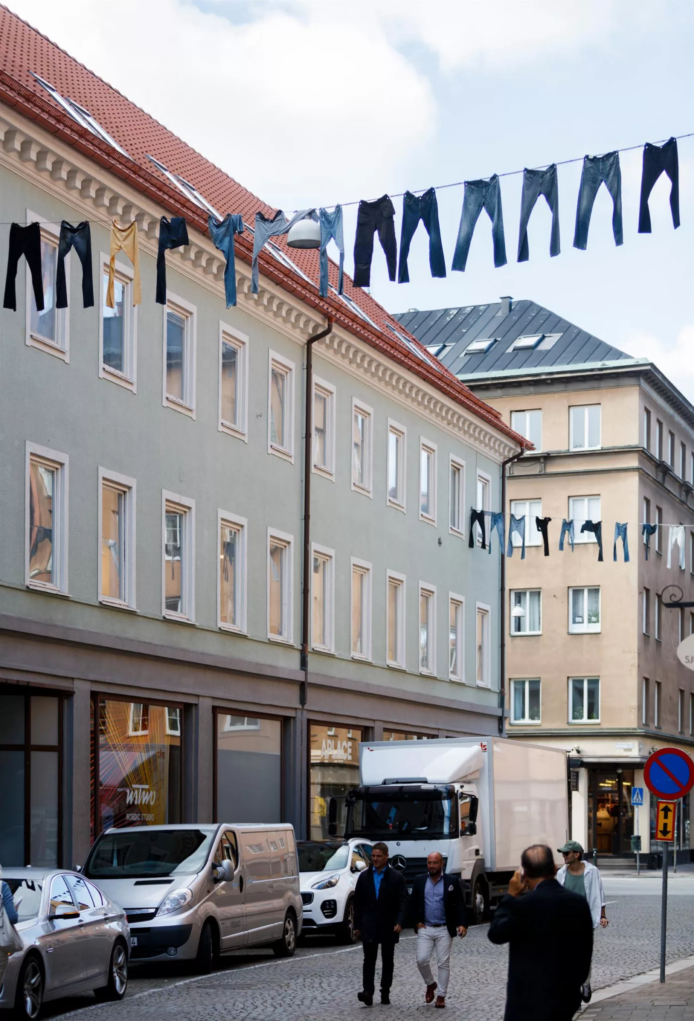 Byggnader med ljusgrön fasad, där jeans hänger på tvättlinor ovanför gatan med skyltfönster och parkerade fordon.