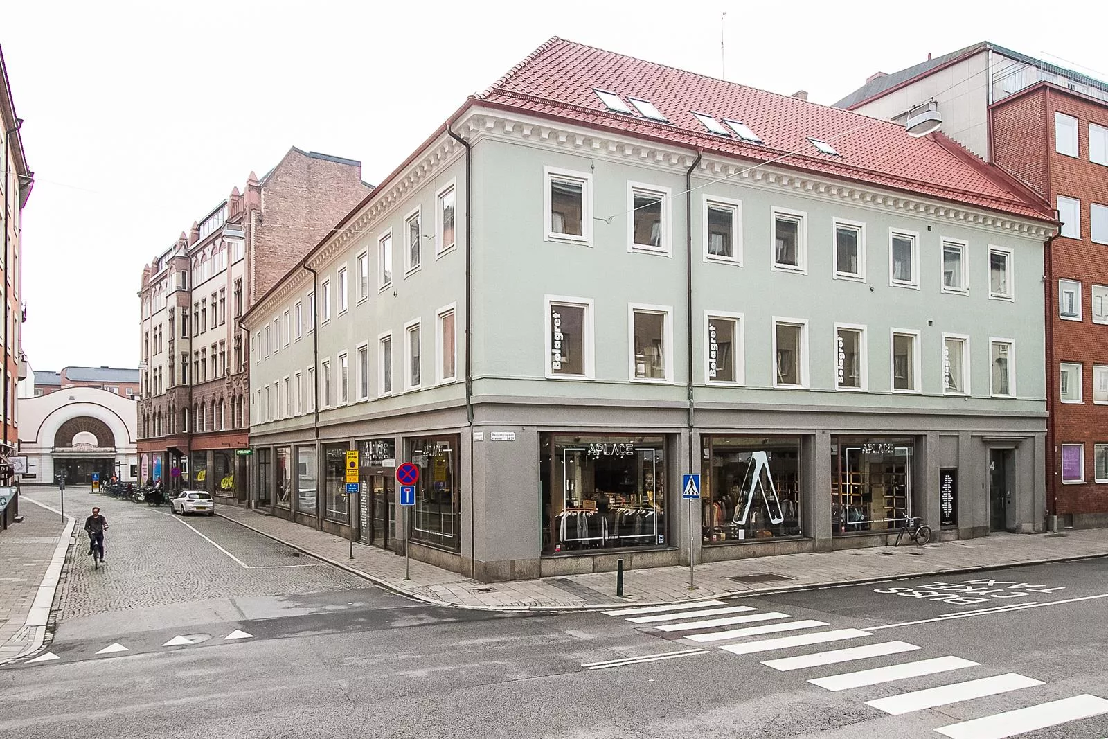 En byggnad med ljusgrön fasad, röda takpannor och stora skyltfönster vid en korsning med övergångsställe.