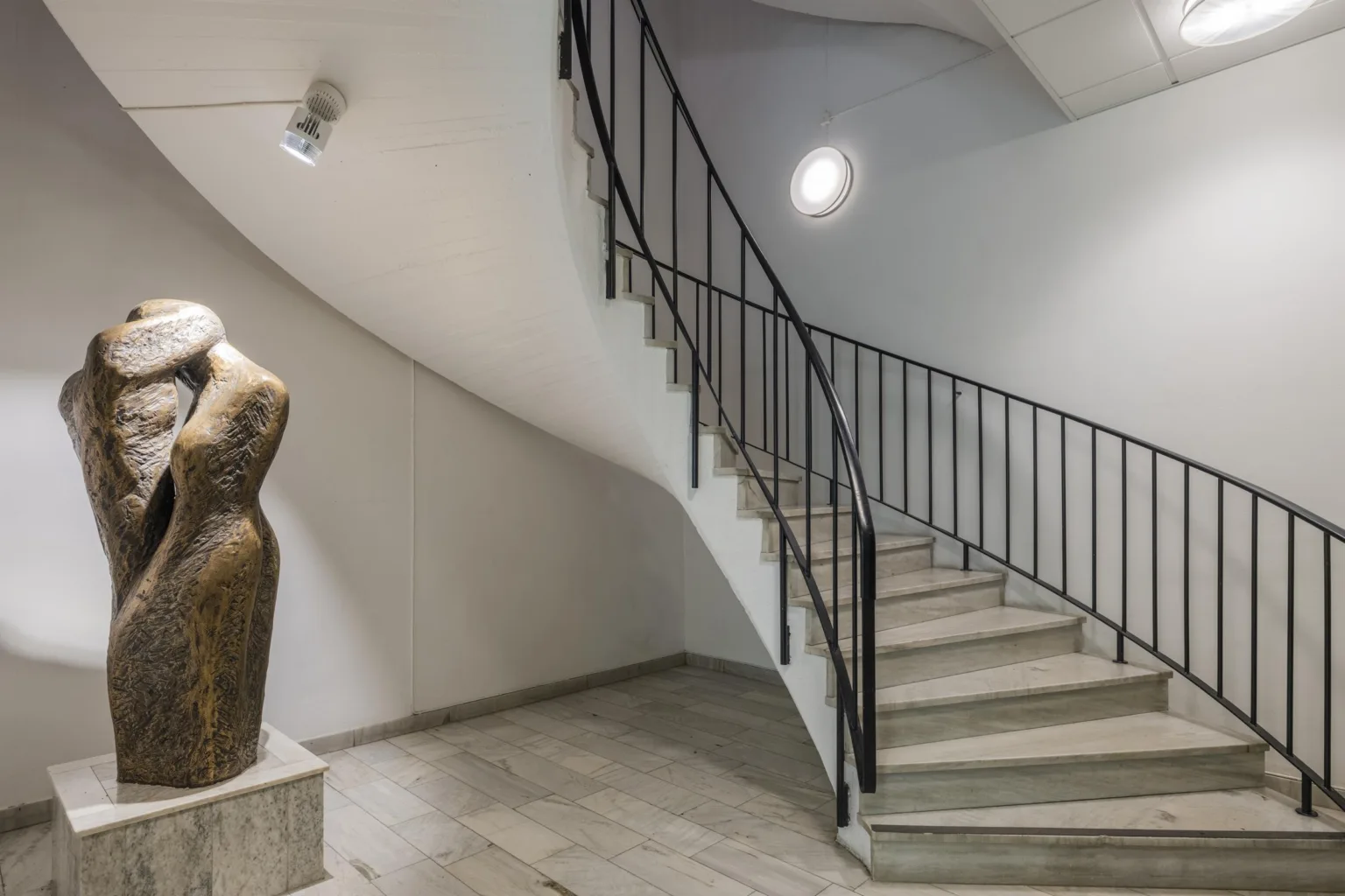 En spiraltrappa med svart räcke i en vit byggnad, bredvid en skulptur på en stenplatta.