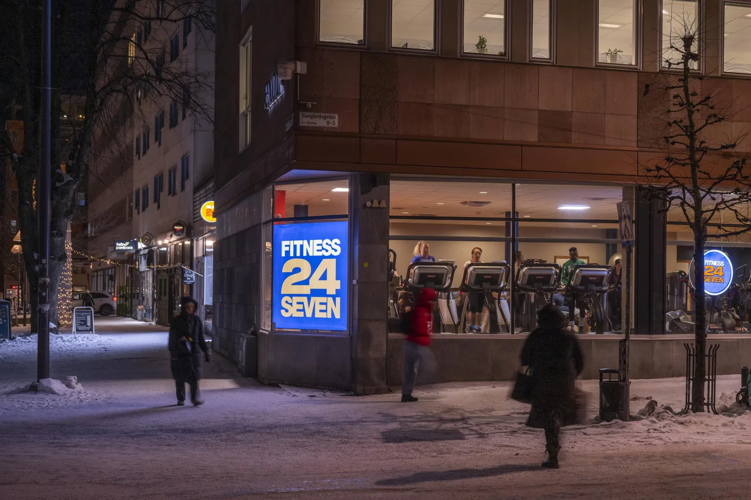 En byggnad med ett gym, Fitness 24 Seven, synligt genom fönstren, omgiven av vinterklädda fotgängare i kvällsbelysning.