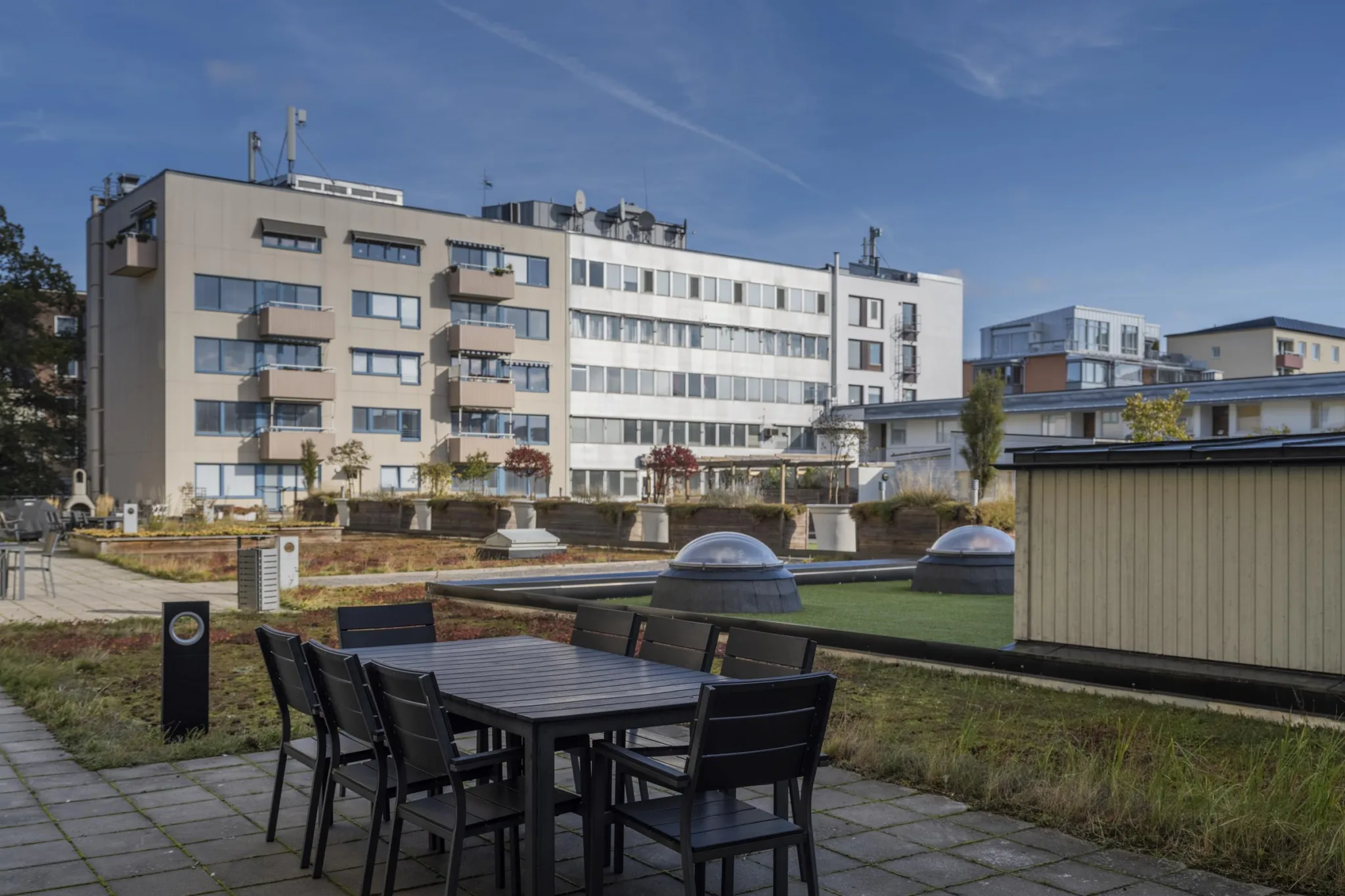 5. En utomhusterrass med bord och stolar, omgiven av gräsyta och byggnader i bakgrunden.