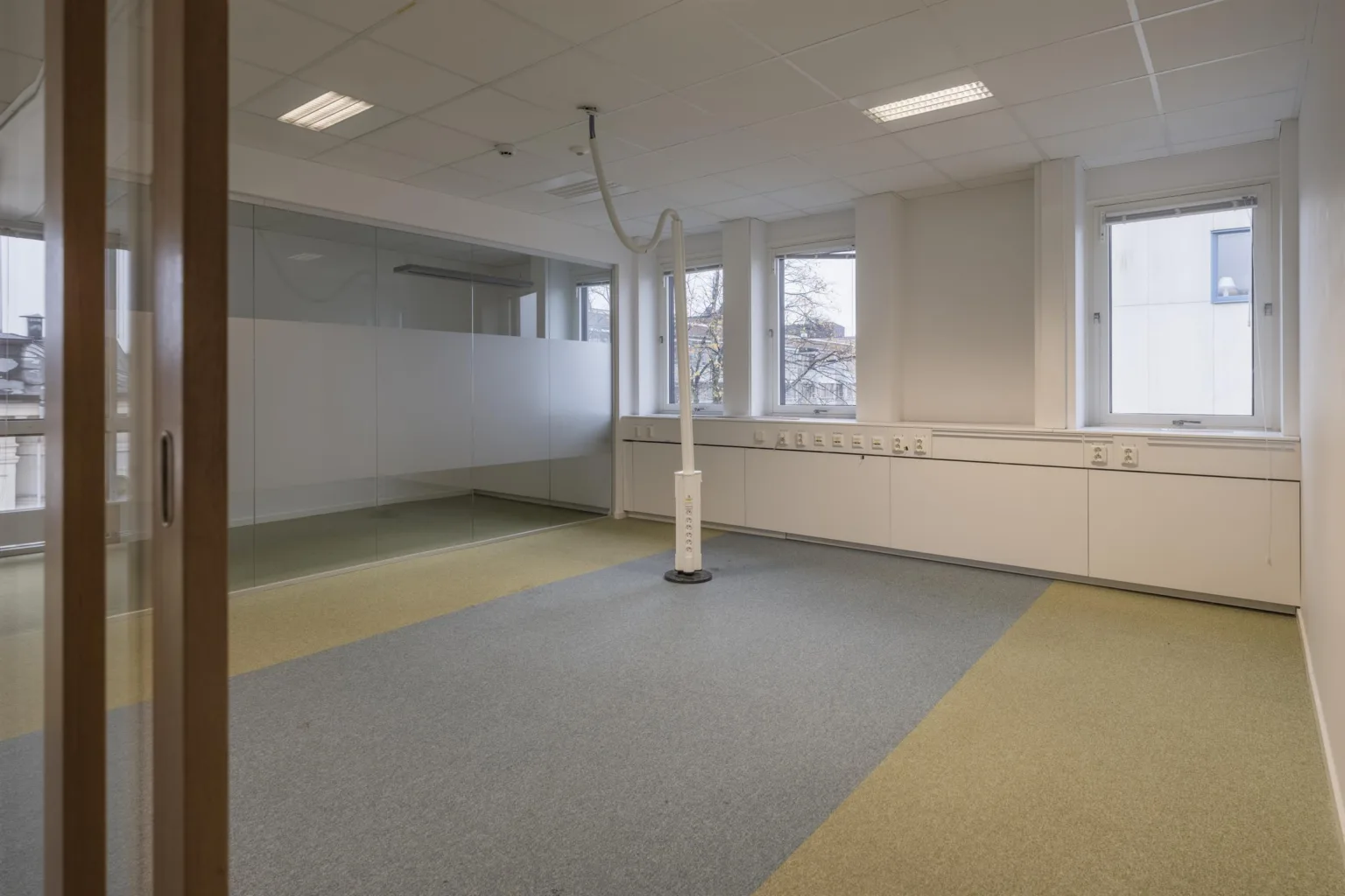 4. En mindre kontorsyta med grönt och blått golv, vita väggar och ett centralt placerat vitt rör.