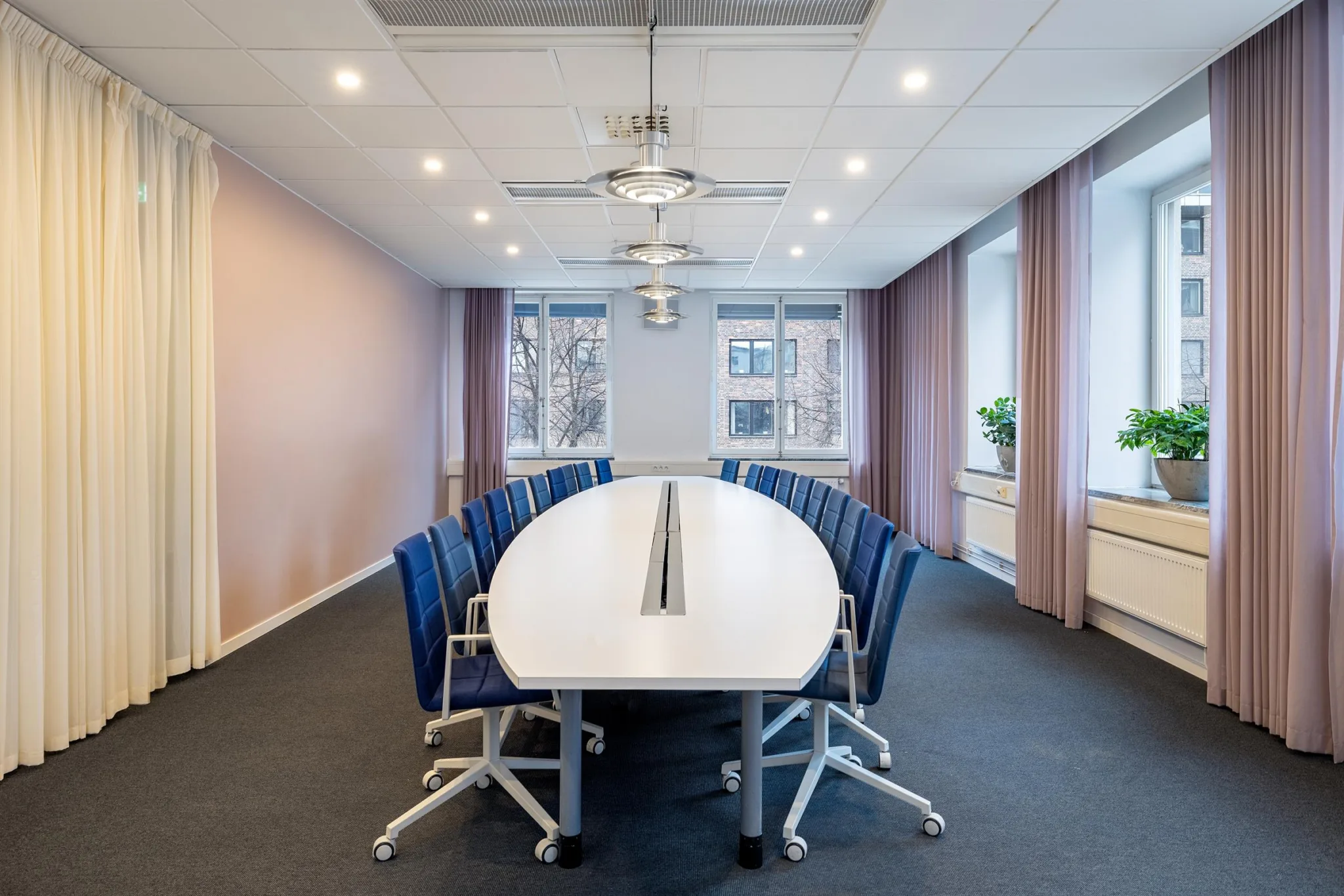 4. Ett konferensrum med ett ovalt vitt bord omgivet av blå stolar och stora fönster med gardiner.