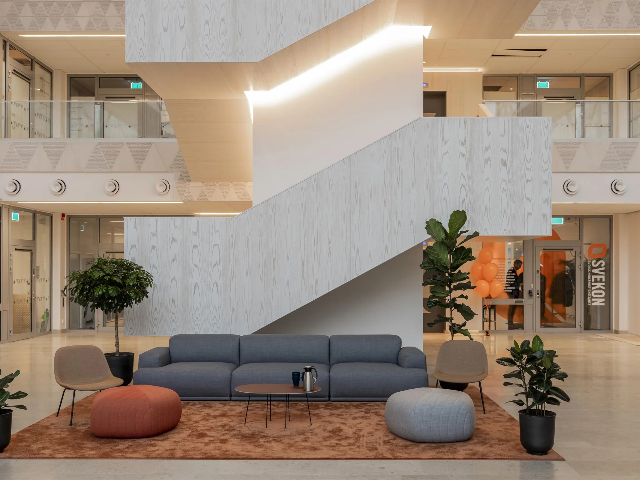 5. Loungeområde med blå soffa, färgglada sittpuffar, växter och en modern trappa i ett luftigt atrium.