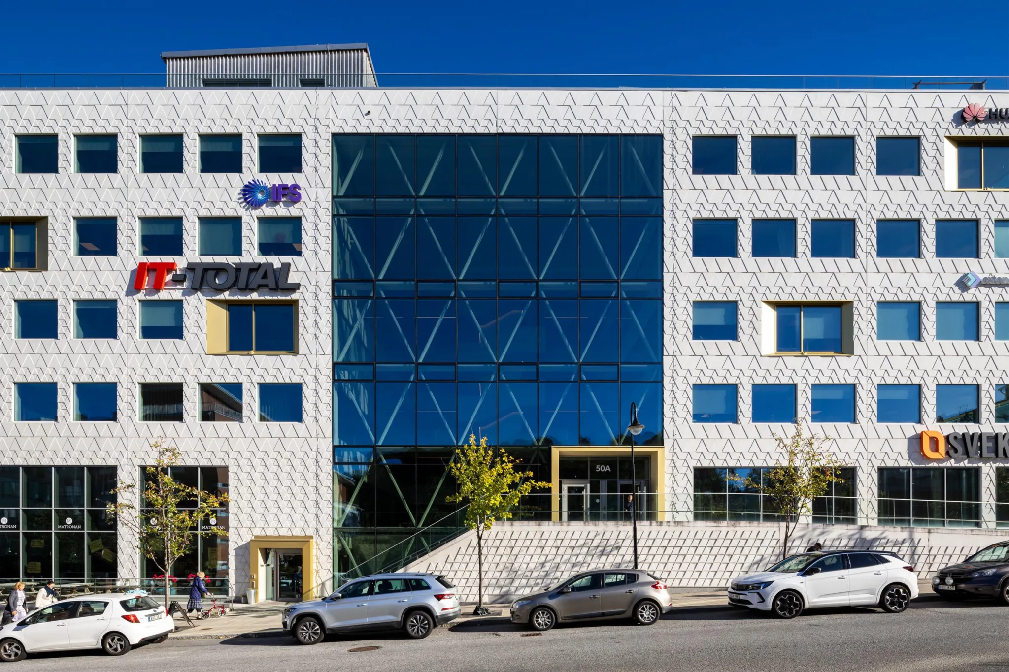 En modern byggnad med glasfasad och vita geometriska mönster, omgiven av parkerade bilar.