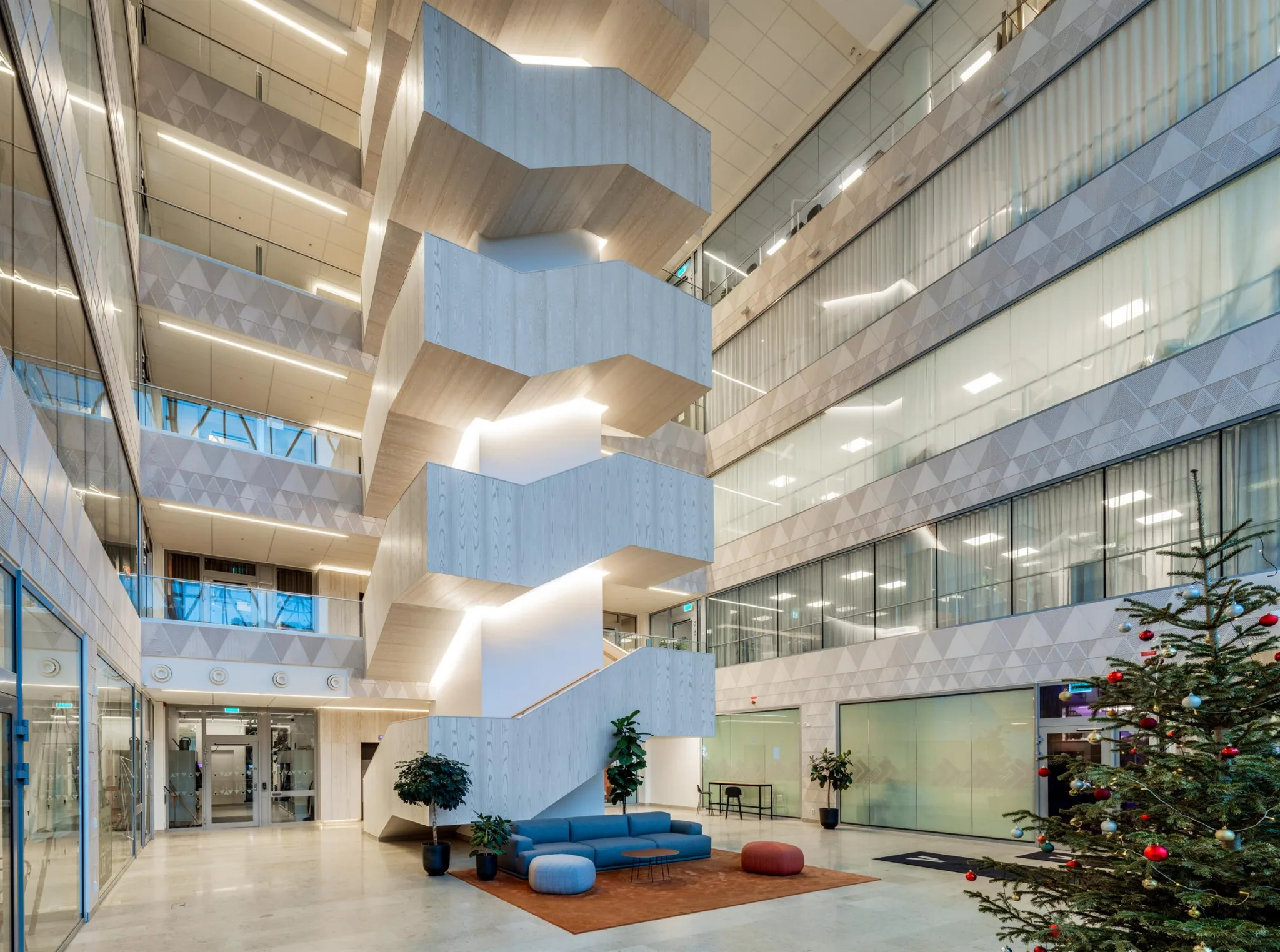 Ett öppet atrium med flera våningar, glasfasader, modern trappa och sittmöbler i mitten.