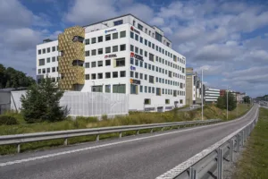 En stor vit byggnad med moderna detaljer och en gul fasaddel, sett från en väg.