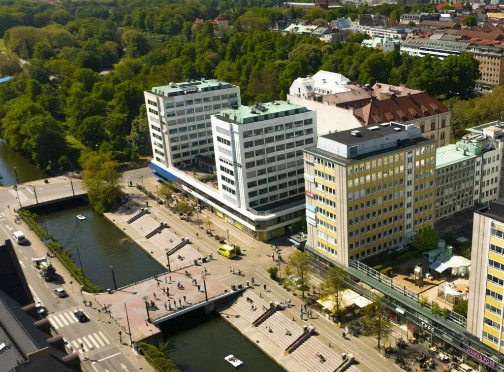 - Flygfoto av flera byggnader och en kanal med trappor och grönska, omgiven av en park och stadsmiljö.