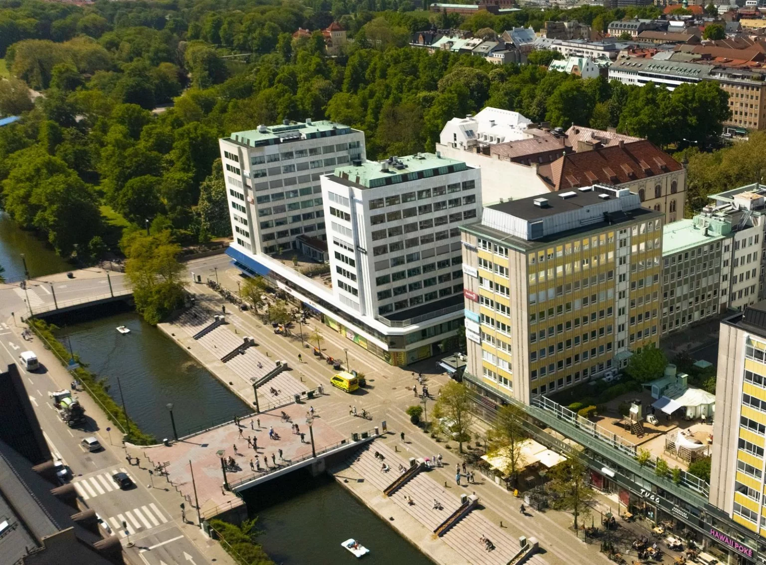 - Flygfoto av flera byggnader och en kanal med trappor och grönska, omgiven av en park och stadsmiljö.