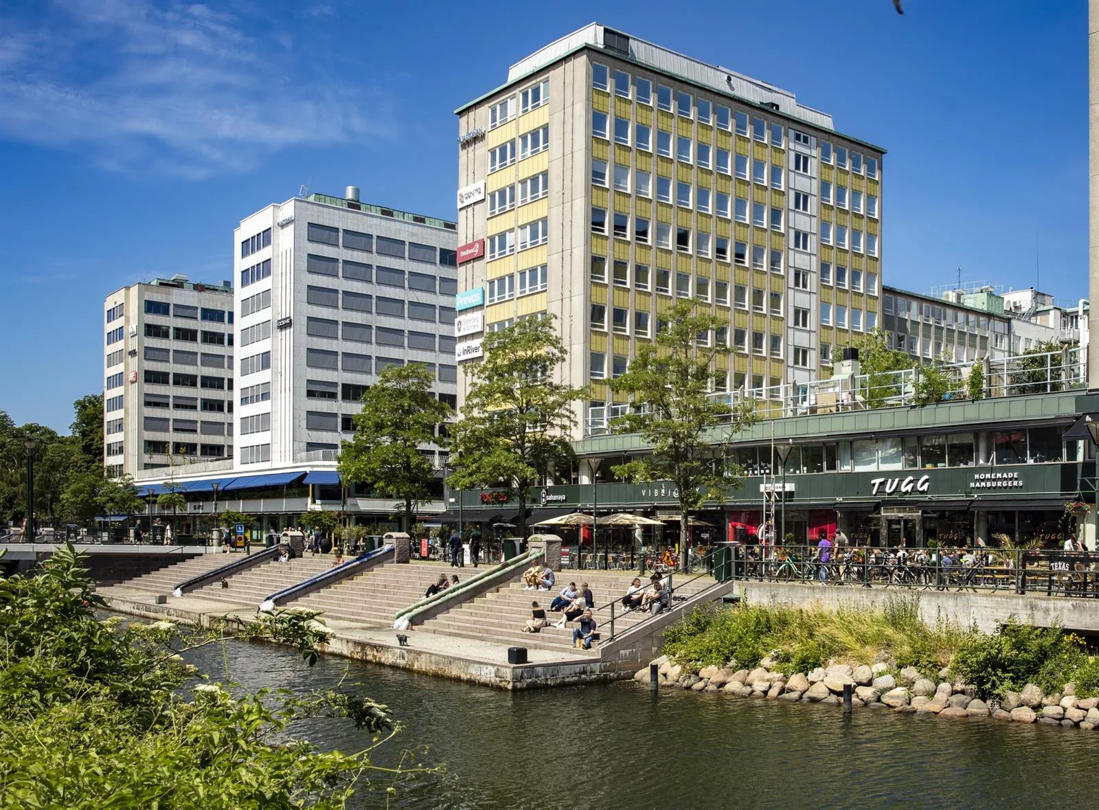 En stadsvy med en kanal, byggnader och människor som sitter vid kajen under en solig dag.