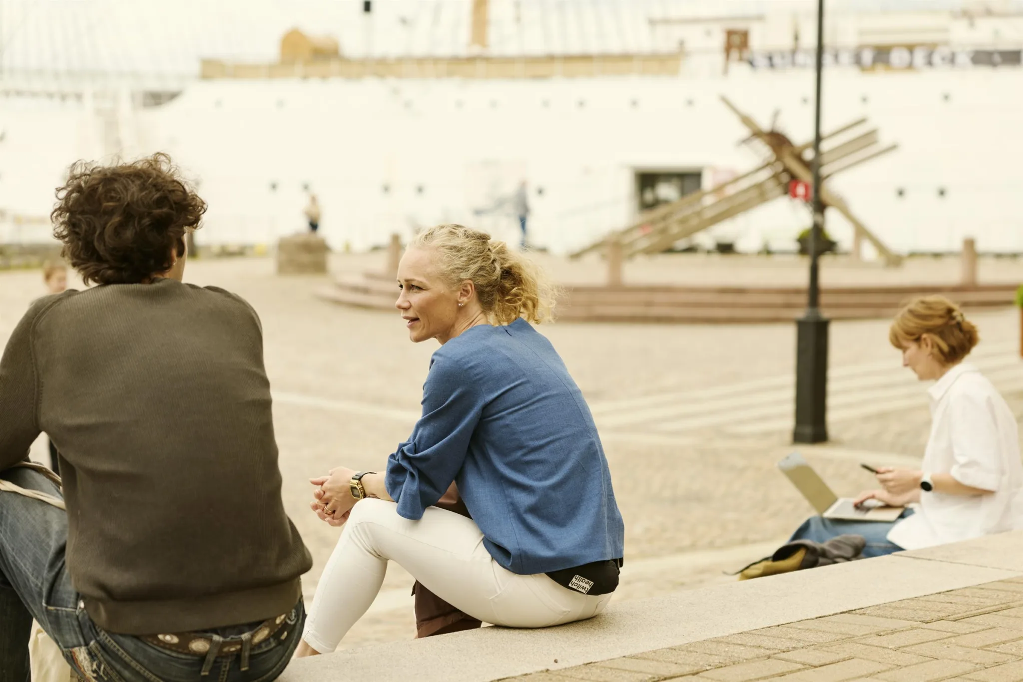 Två personer sitter och pratar på en bänk vid ett torg, med en vit byggnad och trappor i bakgrunden.