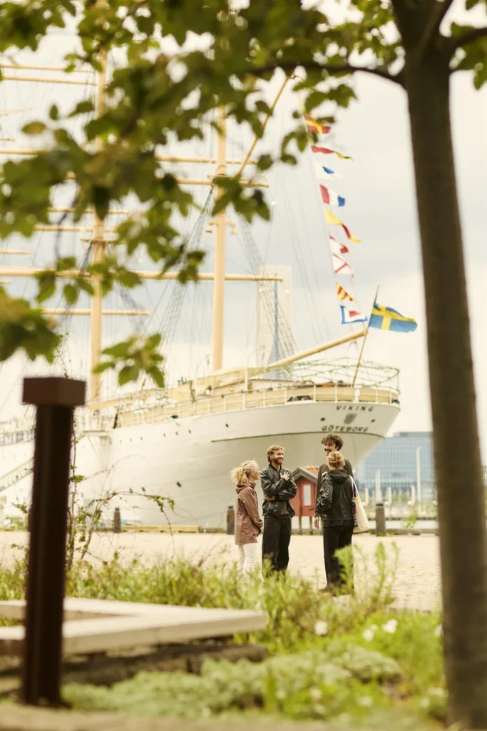 Tre personer står vid en stor båt med svensk flagga och färgglada vimplar.