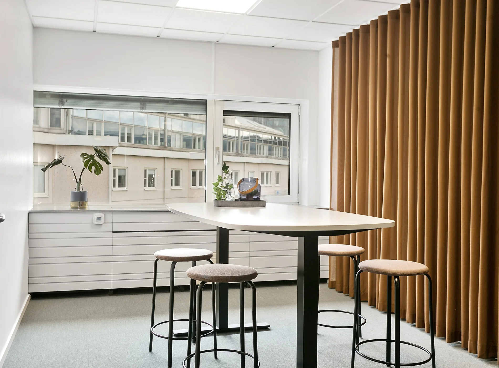 Ett modernt mötesrum med högt bord, fyra pallar, växter och stora fönster som vetter mot en byggnad.