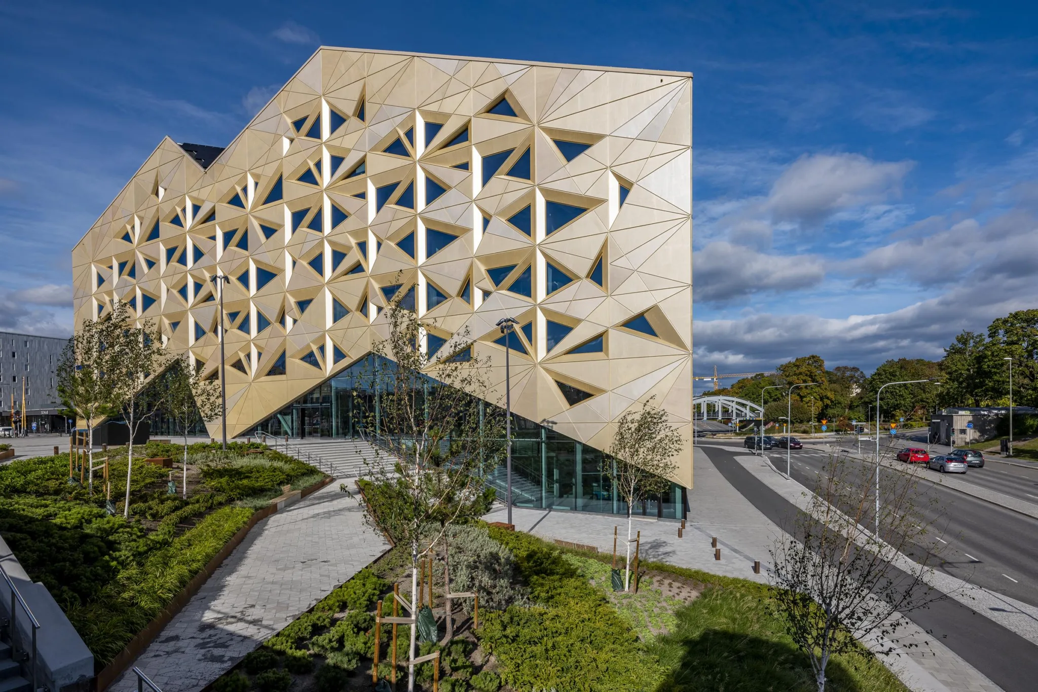 1. En modern byggnad med triangulära fönster och träpaneler, omgiven av grönska och gångvägar under en klar blå himmel.