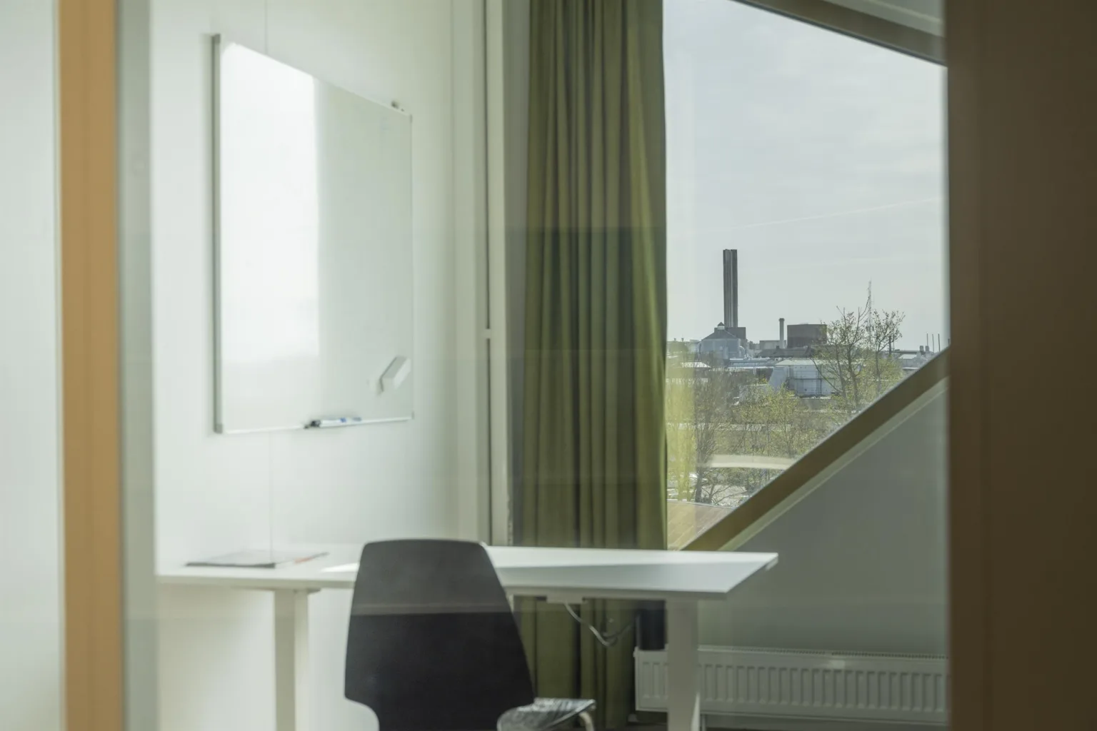 2. Ett ljust kontorsrum med skrivbord, svart stol, grön gardin och utsikt över industriskorsten genom ett geometriskt fönster.