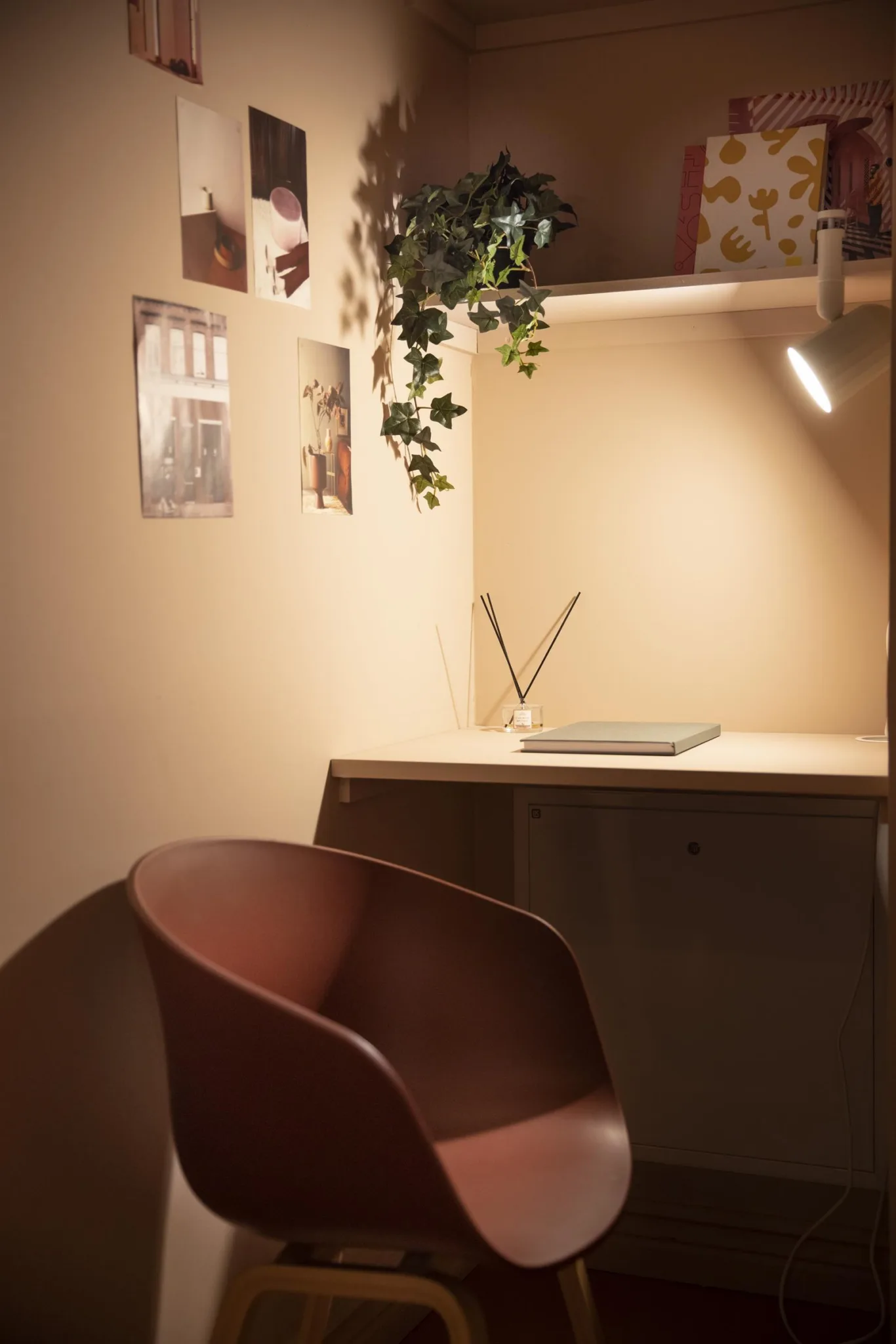 1. Ett litet skrivbord med en laptop, väggdekorationer och en hängande växt, bredvid en brun stol under varm belysning.