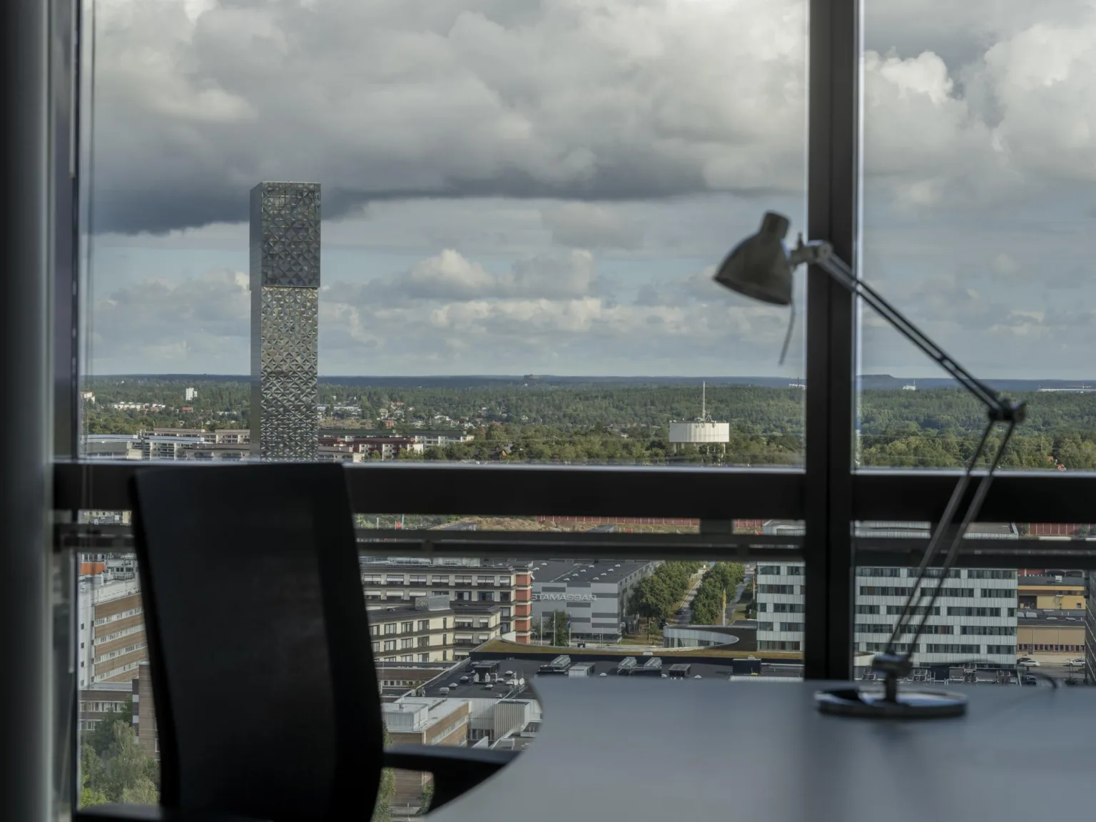 Ett kontorsrum med en skrivbordslampa och stol framför stora fönster med utsikt över en stad.