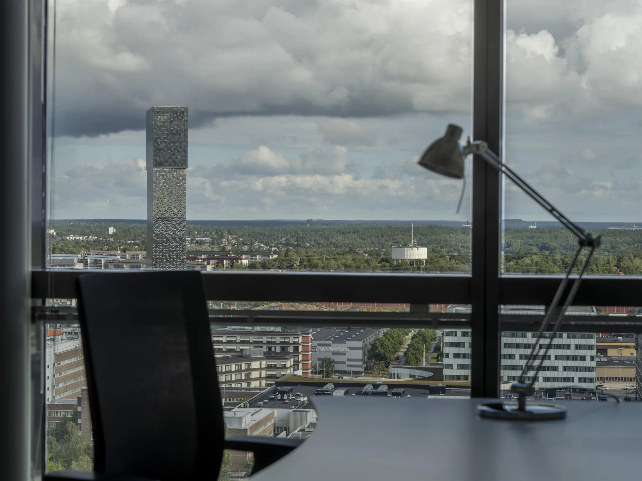 Ett kontorsrum med en skrivbordslampa och stol framför stora fönster med utsikt över en stad.