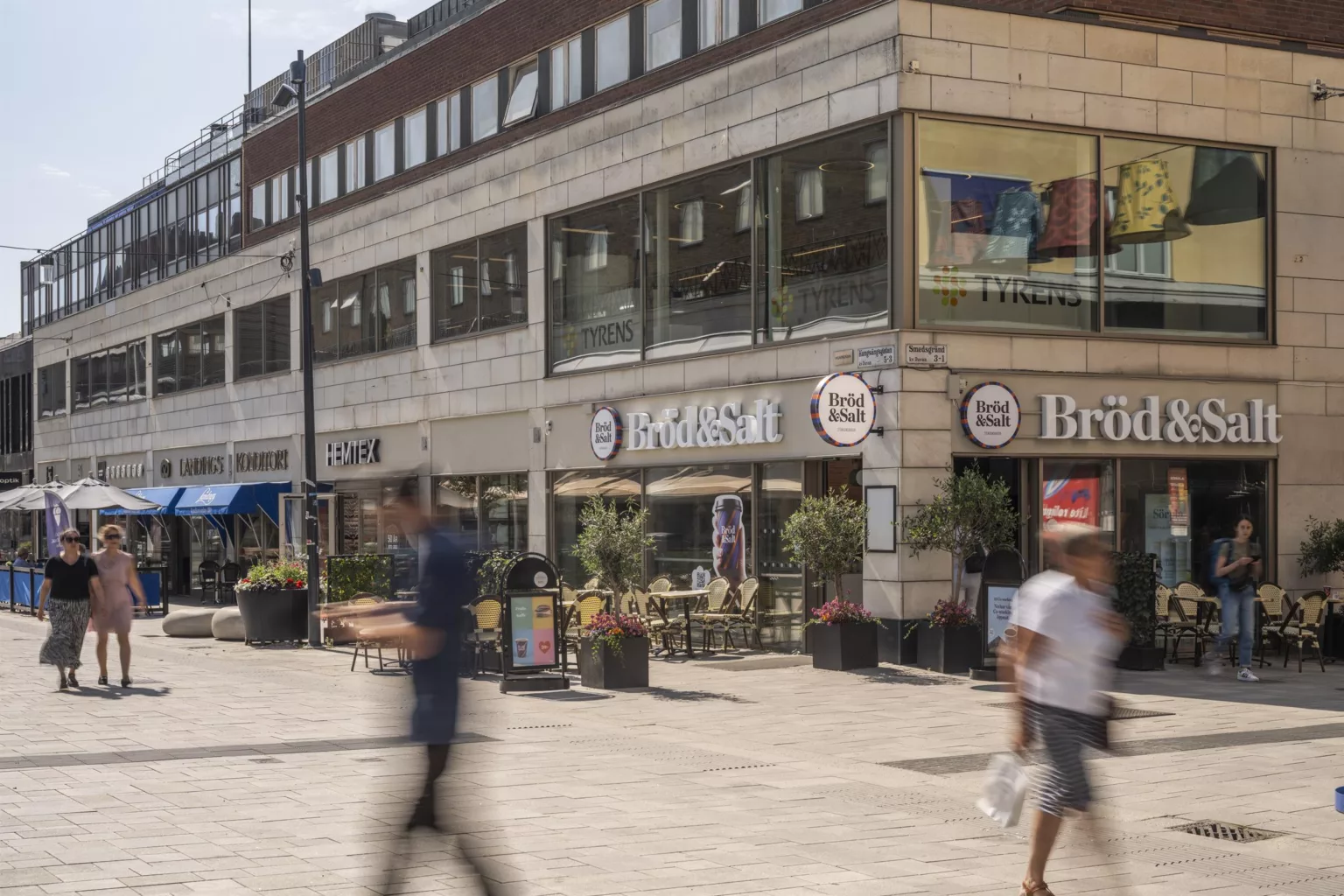 En stadsmiljö med affärer och kaféer, folk går förbi på en bred trottoar.