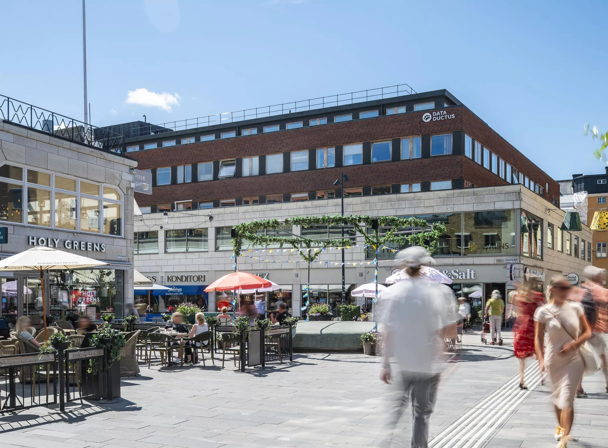 En utomhusservering med parasoller och bord, människor rör sig genom ett öppet torg under en klar himmel.
