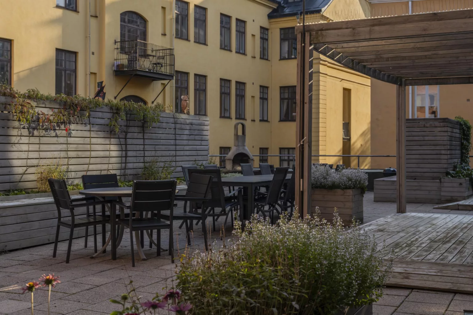En innergård med bord och stolar omgivna av växter, med en gul byggnad i bakgrunden.
