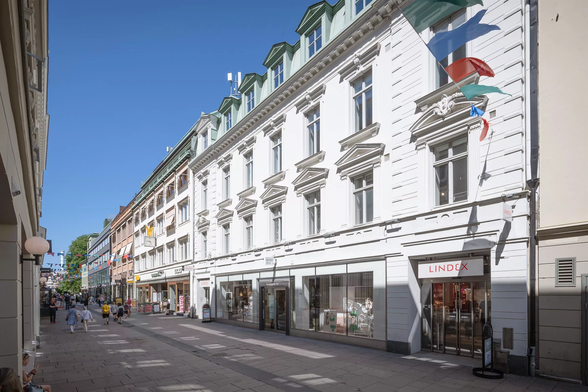 En vit byggnad med flera våningar, stora fönster, och affärer på bottenplan längs en stadsgata under klarblå himmel.