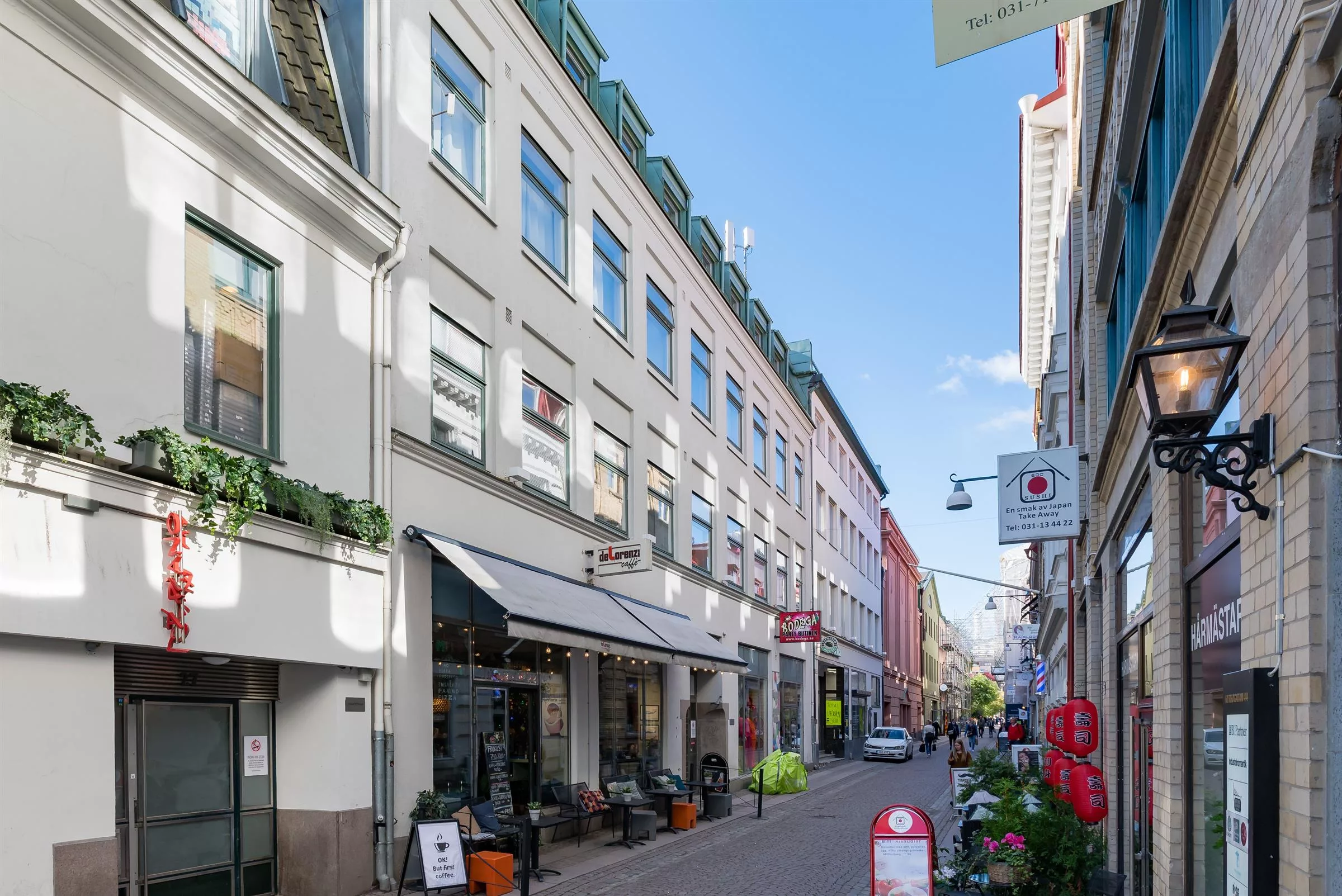 En smal stadsgata med flervåningshus på båda sidor, affärer, kaféer och skyltar under en solig himmel.