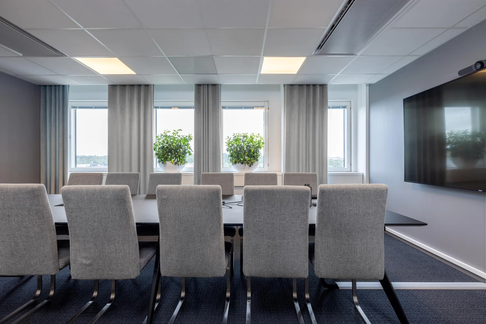 Ett konferensrum med ett långt bord, grå stolar och stora fönster med gardiner och gröna växter.