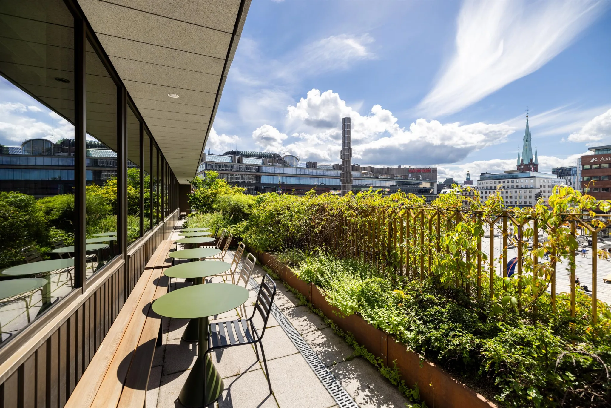 En terrass med gröna bord och stolar, omgiven av växter och med utsikt över en stadsmiljö under en blå himmel med moln.
