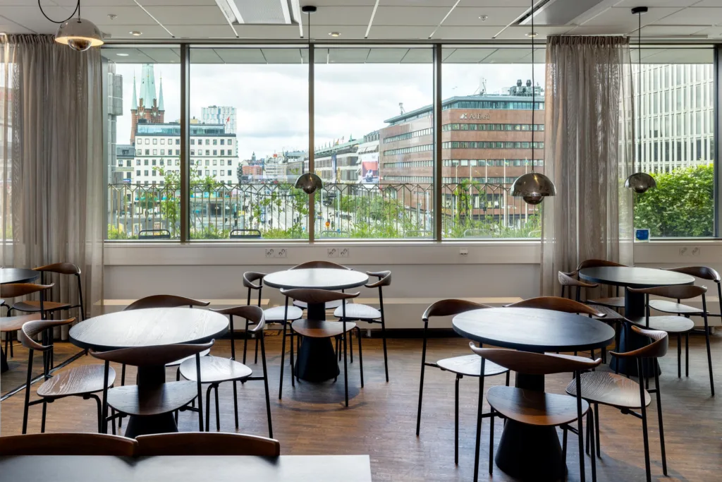En matsal med runda bord och stolar, stora fönster med utsikt över en stadsmiljö med byggnader och kyrktorn.