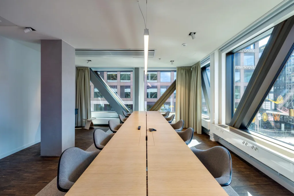 Ett konferensrum med ett långt bord, grå stolar och stora fönster med gardiner.