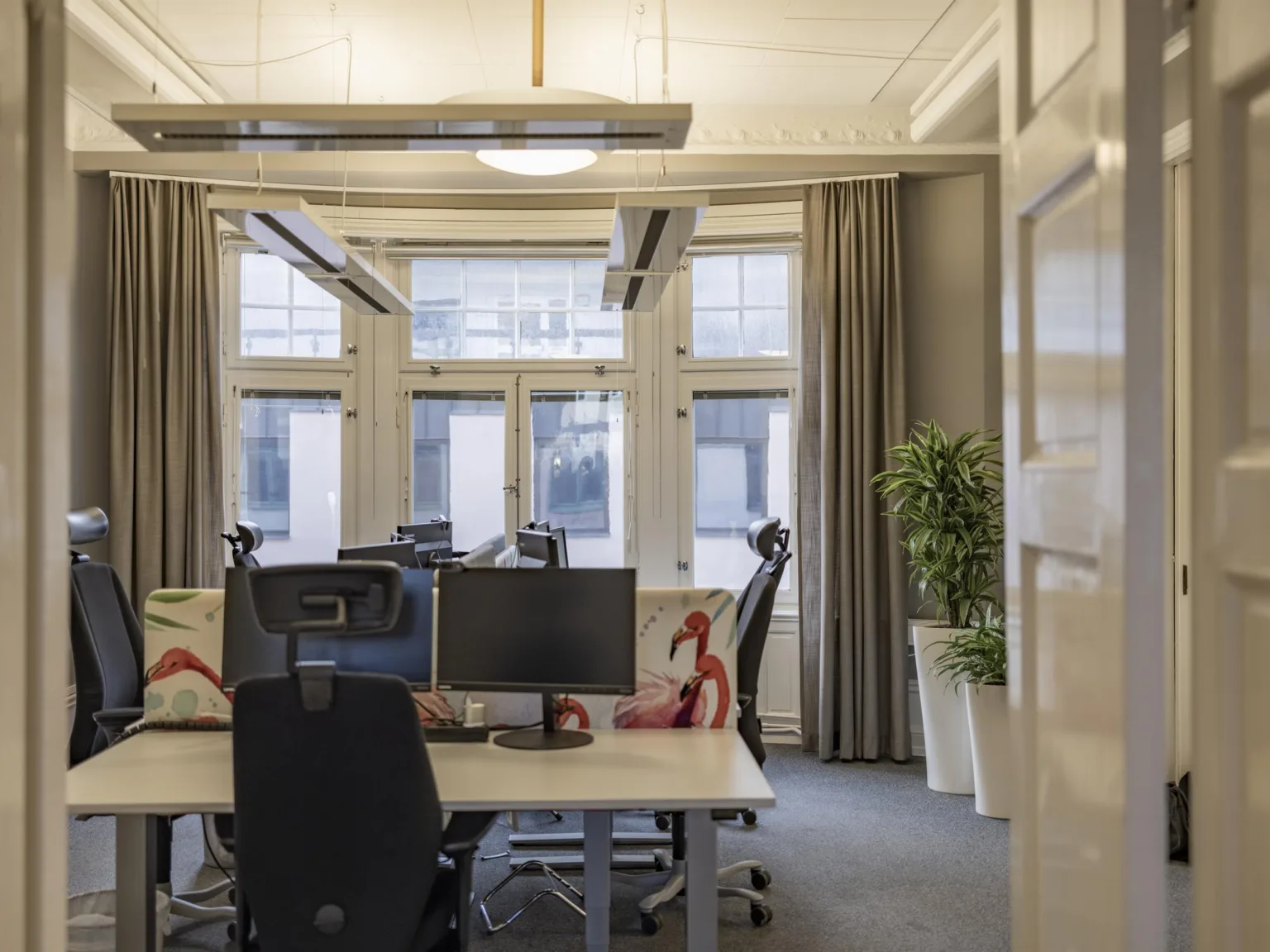 Kontorsrum med skrivbord, datorskärmar, stolar och stora fönster med gardiner.