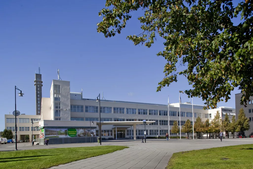 2. En modern byggnad med flera våningar och stora fönster, omgiven av grönska och en gångväg under en klar blå himmel.