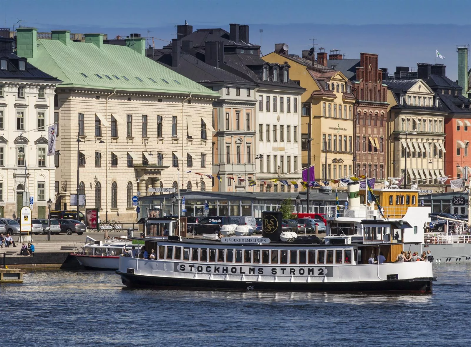 En vit turistbåt med texten "Stockholm Ström" på vattnet, framför färgglada historiska byggnader.