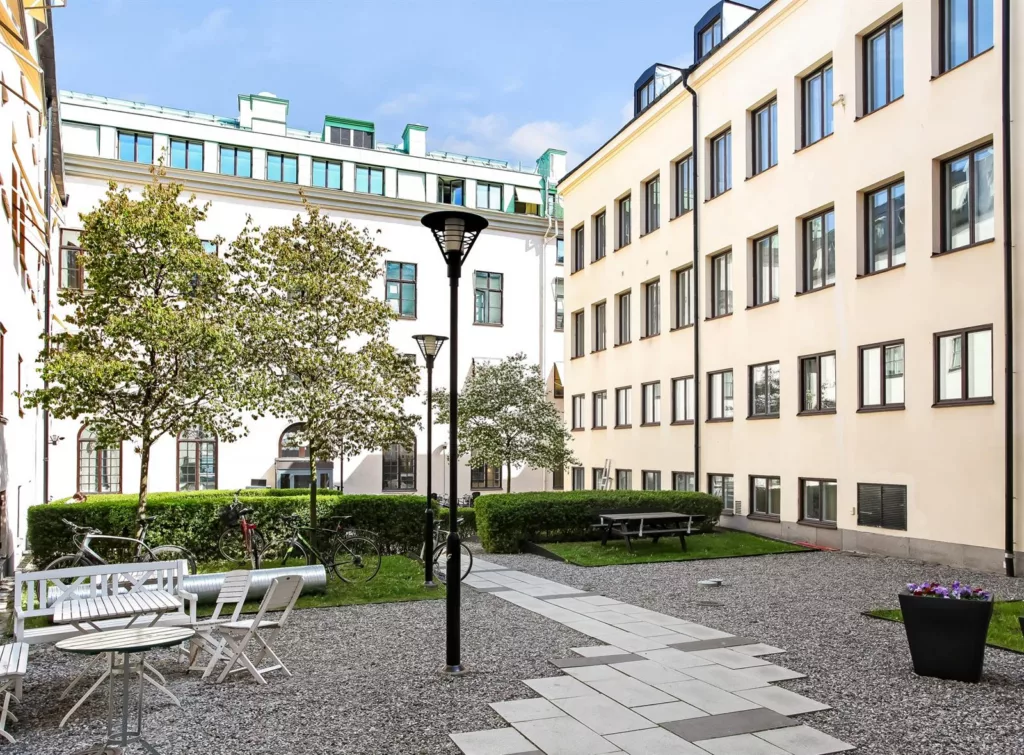 En innergård med träd, buskar, sittmöbler och belysningsstolpar, omgiven av ljusa byggnader.