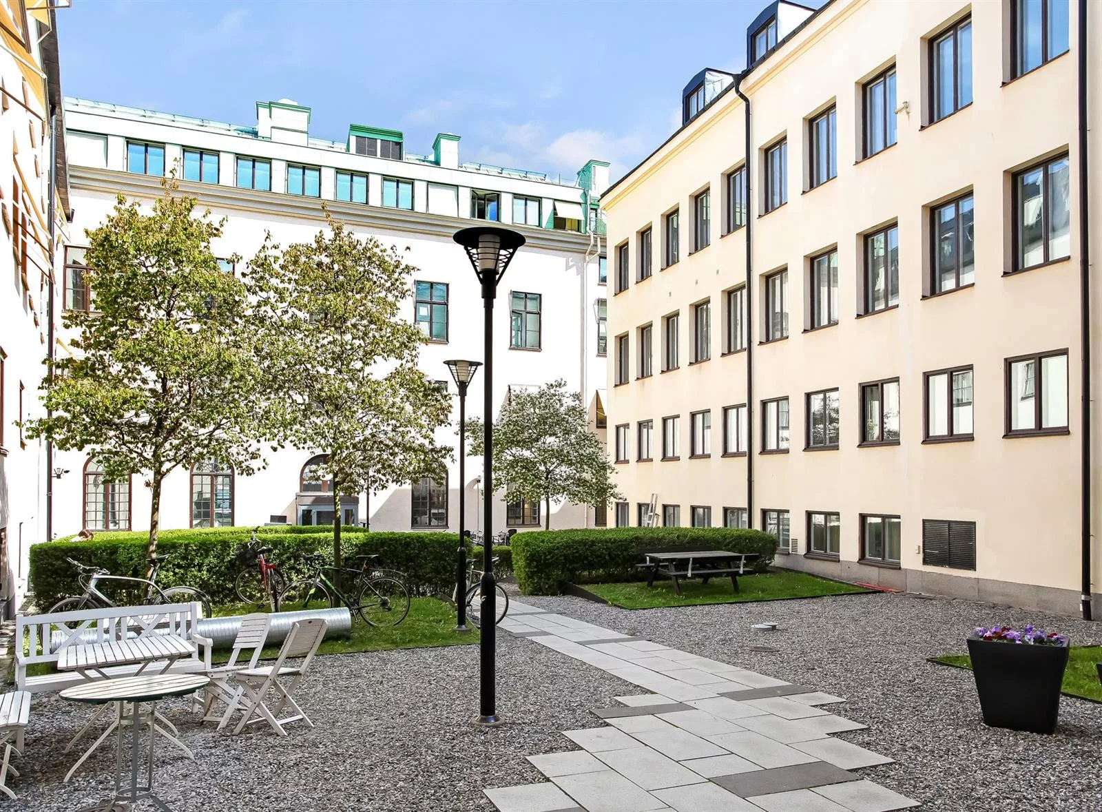 En innergård med träd, buskar, sittmöbler och belysningsstolpar, omgiven av ljusa byggnader.
