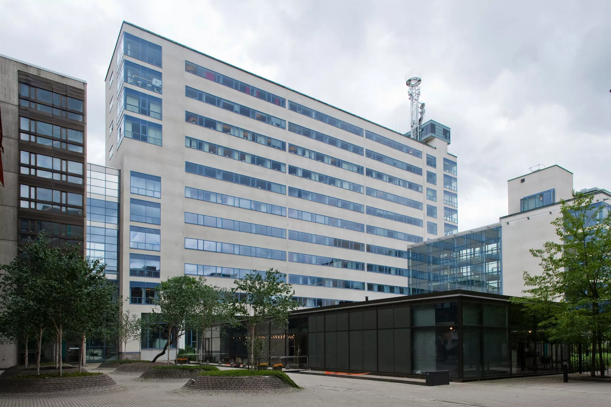 En hög byggnad med glasfasad, omgiven av träd och mindre byggnader.